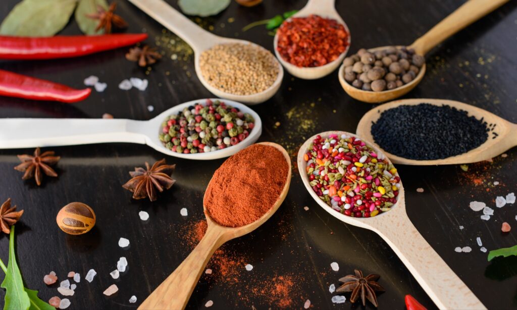 Spoonfuls of spices and herbs arranged in a circle on a table. Extra spices and herbs are scattered around the table.