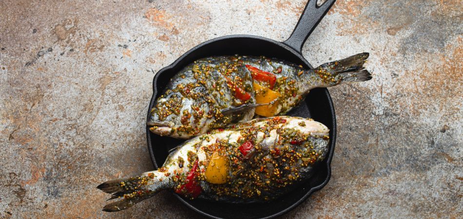 Two grilled fish in a skillet. There is also a salad and cutlery on the table.